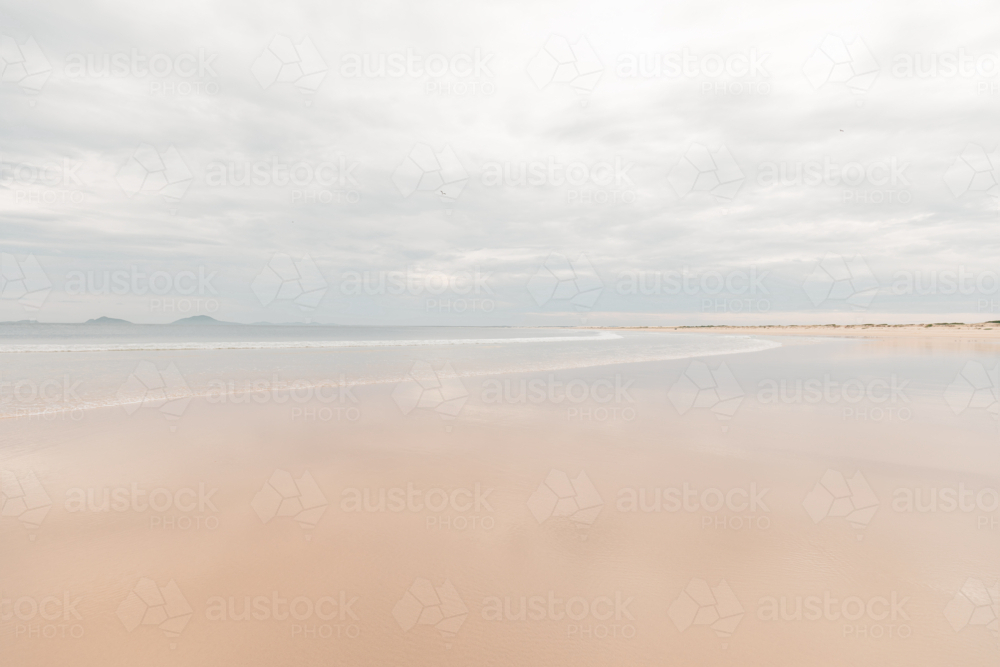 Expansive sandy beach with gentle waves under a cloudy sky and distant hills - Australian Stock Image