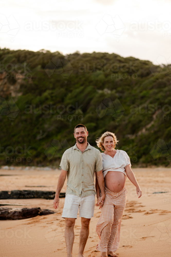 Expecting couple walking hand in hand on a beach during sunset - Australian Stock Image