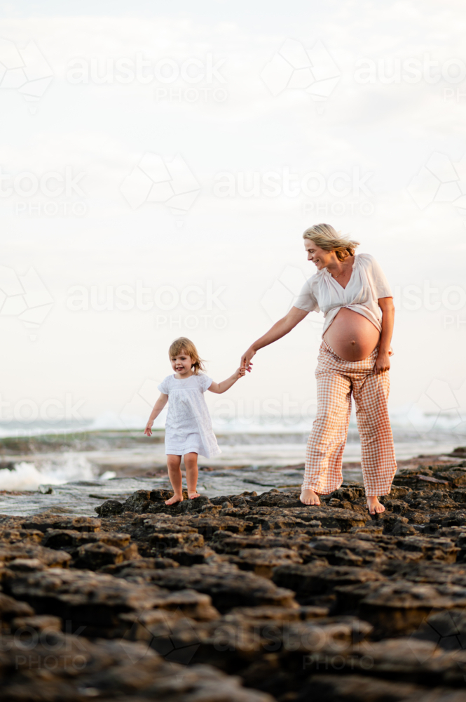 Expecting mother and daughter walking hand in hand on rocky beach at sunset - Australian Stock Image
