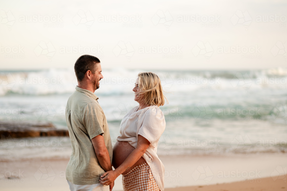 Expecting parents smiling at each other during a sunset at the beach - Australian Stock Image