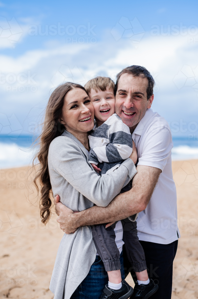 Family embraces happily on the beach, with a child laughing between parents by the ocean - Australian Stock Image