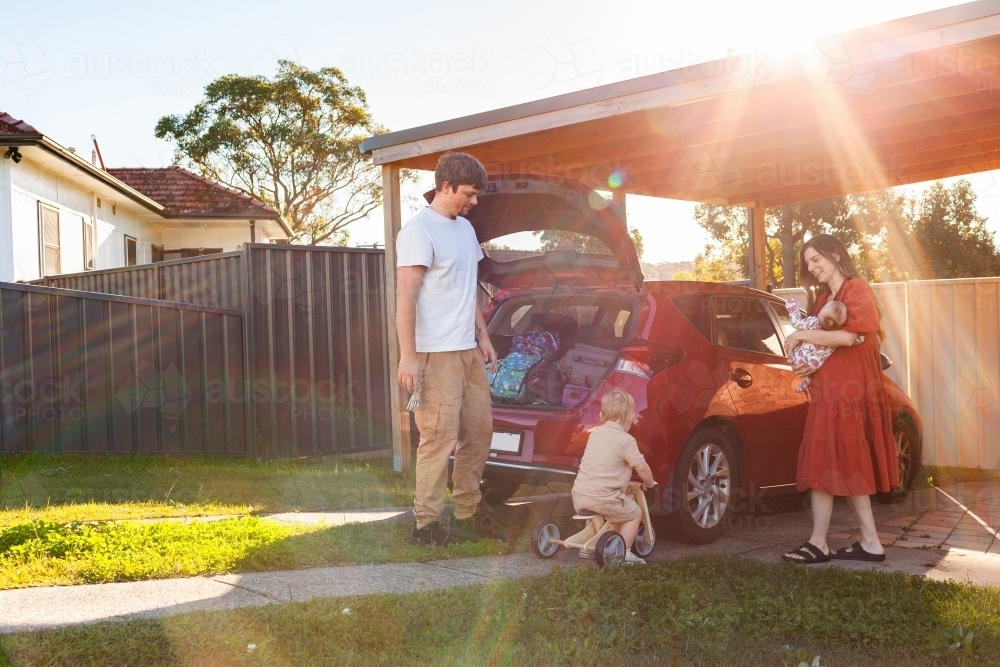 Family of four packing car boot with luggage for holidays with toddler riding away on trike - Australian Stock Image