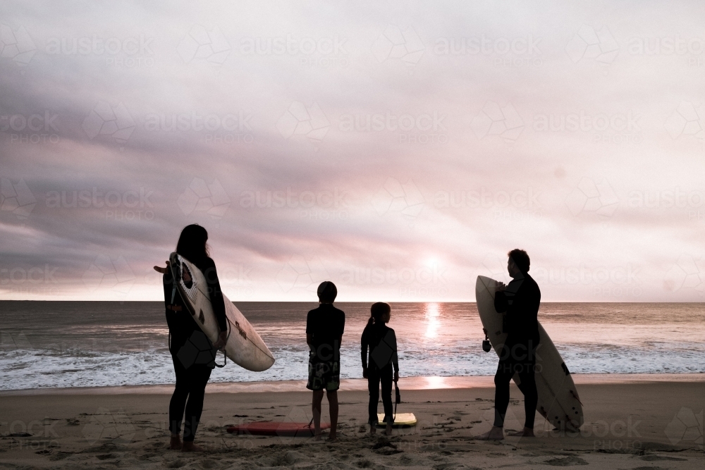 Family of four standing on beach in silhouette holding surfboards with moody sunset - Australian Stock Image