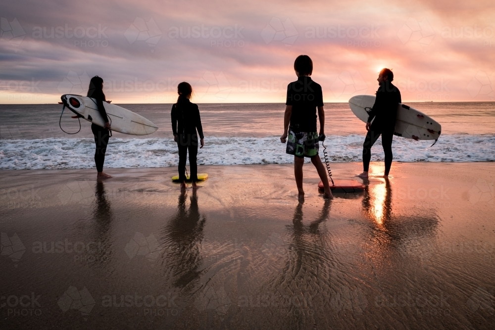 Family of four standing on beach in silhouette holding surfboards with moody  sunset - Australian Stock Image