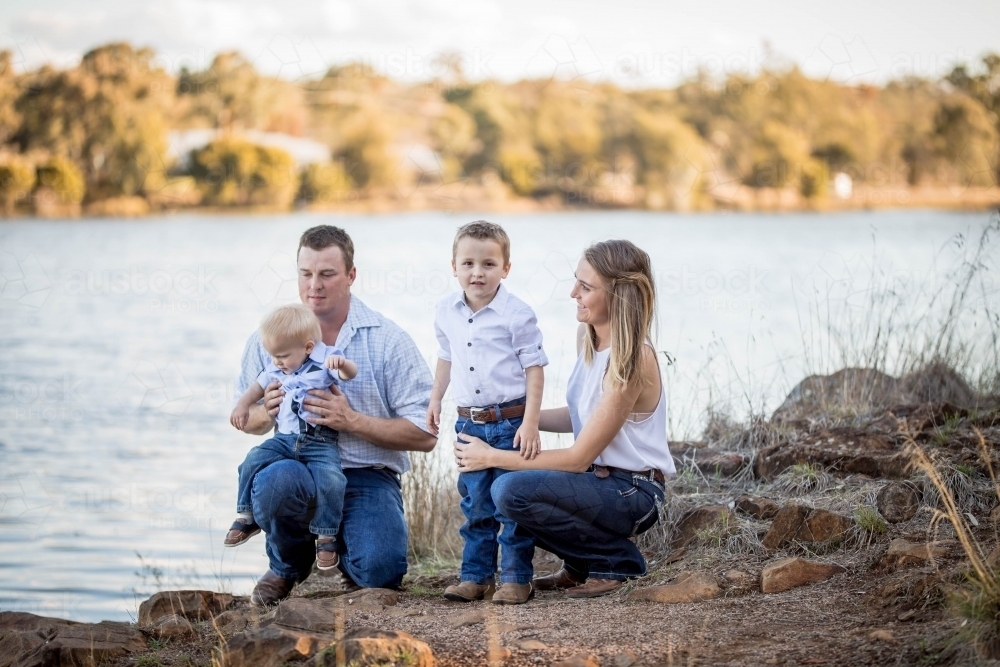Family of husband and wife with two sons kneeling down near water - Australian Stock Image