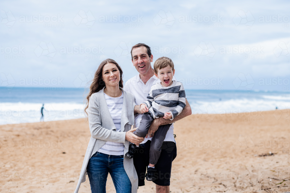 Family of three happy on a sandy beach with ocean waves in the background - Australian Stock Image