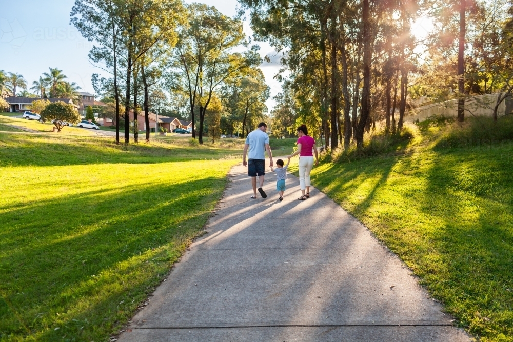 Family of three holding hands with toddler on suburban walk in afternoon light - Australian Stock Image
