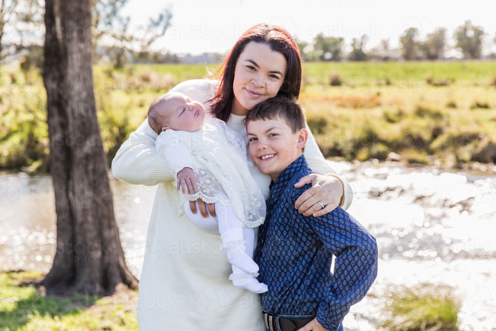 Family of three mum, son and newborn baby girl cuddling and happy near river - Australian Stock Image