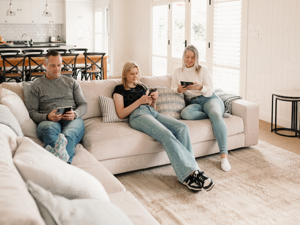 Family of three sitting comfortably on the couch while on their phones - Australian Stock Image