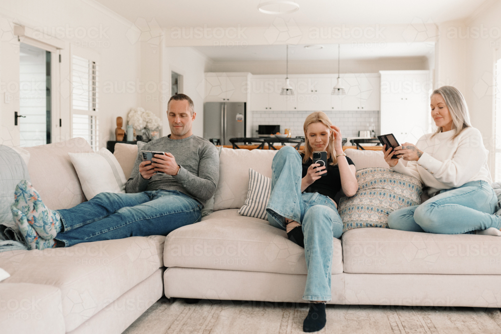 Family of three sitting comfortably on the couch while on their phones - Australian Stock Image