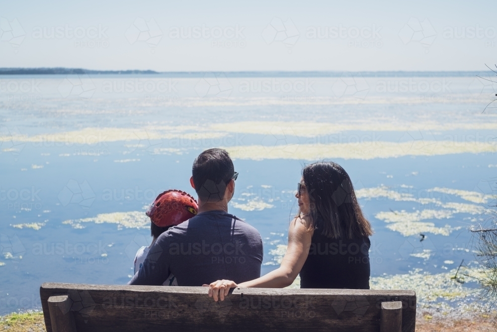 Family of three sitting on bench looking out over water - Australian Stock Image