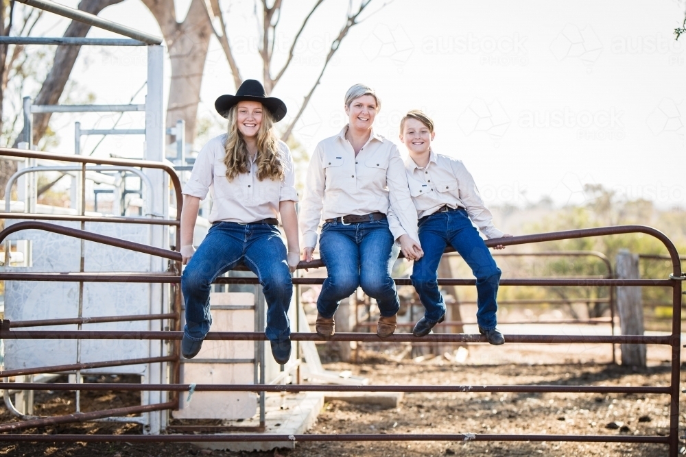 Family of three sitting on gate in stock yards on farm in drought - Australian Stock Image