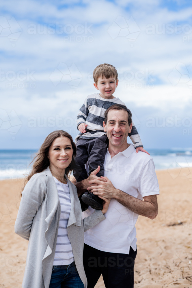 Family of three smiles happily together on a beach with the ocean behind them - Australian Stock Image