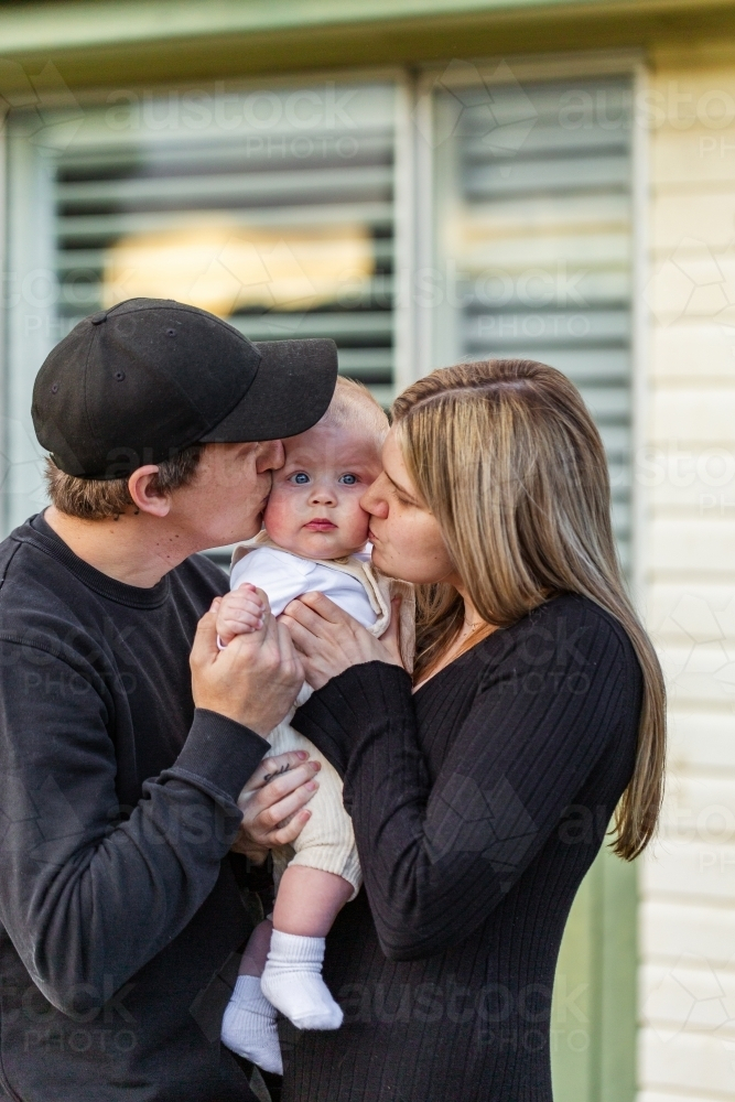 Family of three with parents kissing child outside of home at sunset - Australian Stock Image