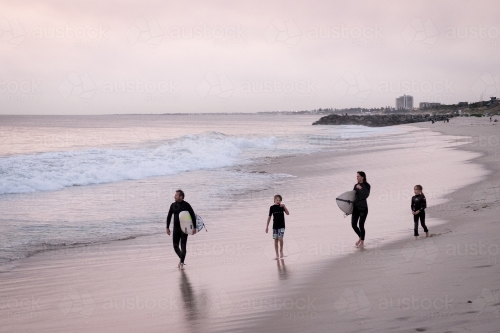 Family of two adults and two children walking along the beach carrying surfboards dressed in wetsuit - Australian Stock Image