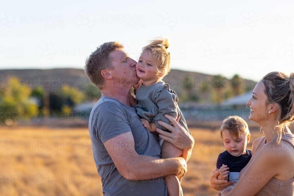 family outside with dad kissing little girl and mum and son looking on - Australian Stock Image