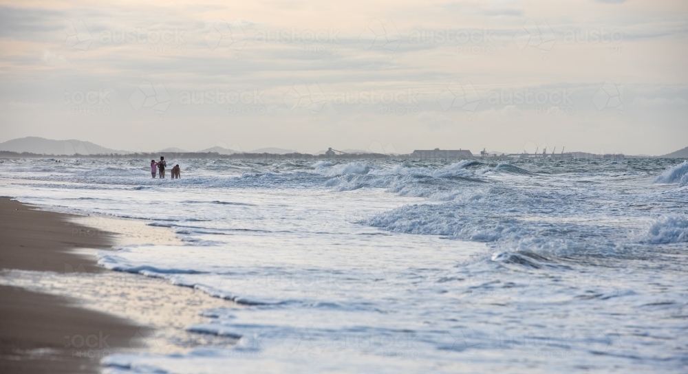 Family playing in the waves on Tannum Sands beach with industry in the background, Gladstone Region, - Australian Stock Image