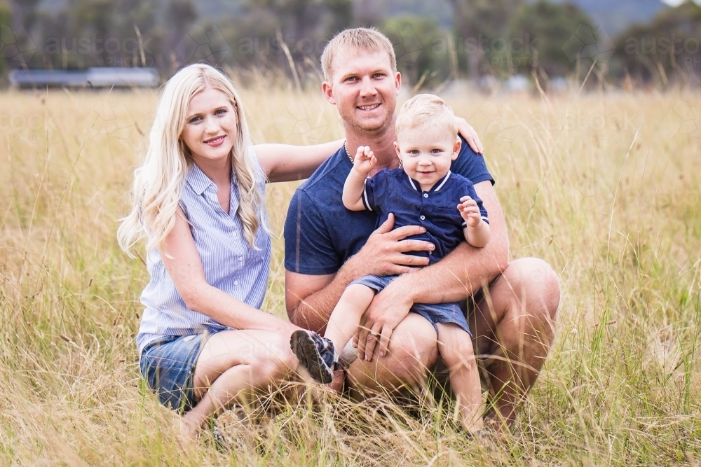 Family sitting in long grass in paddock on farm smiling - Australian Stock Image