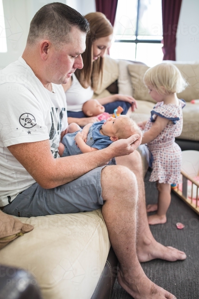 Family sitting on lounge mother playing with daughter father holding newborn baby boy - Australian Stock Image