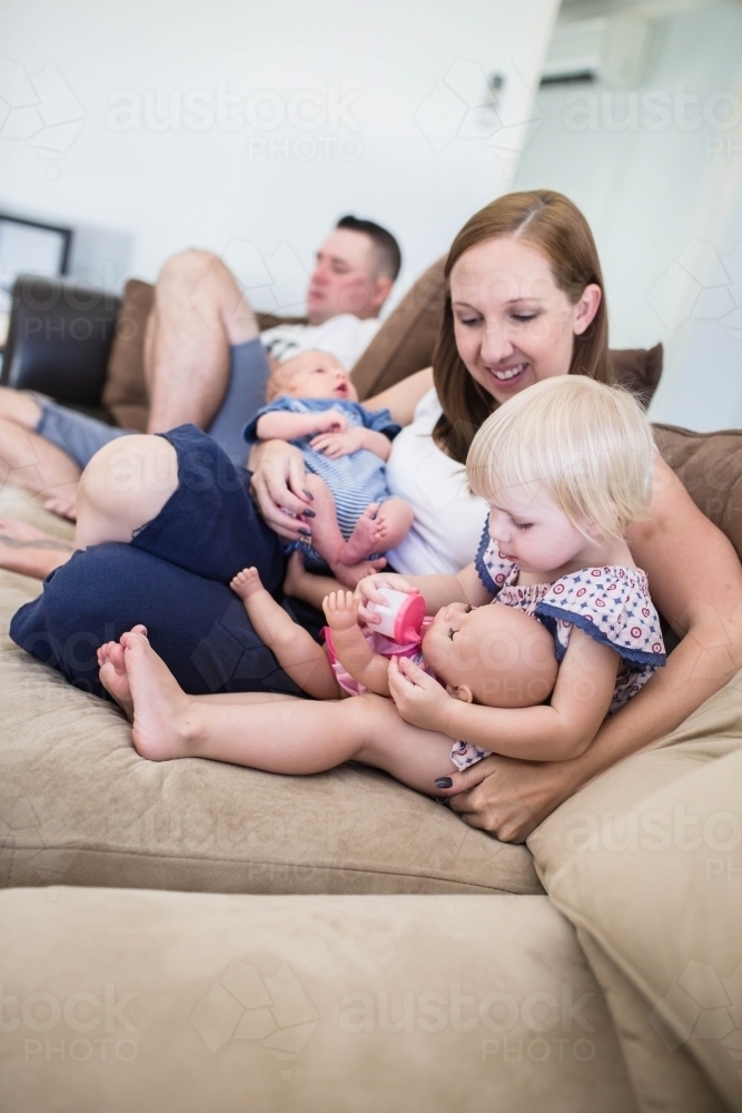 Family sitting on lounge mum holding newborn baby boy while playing with daughter and doll - Australian Stock Image