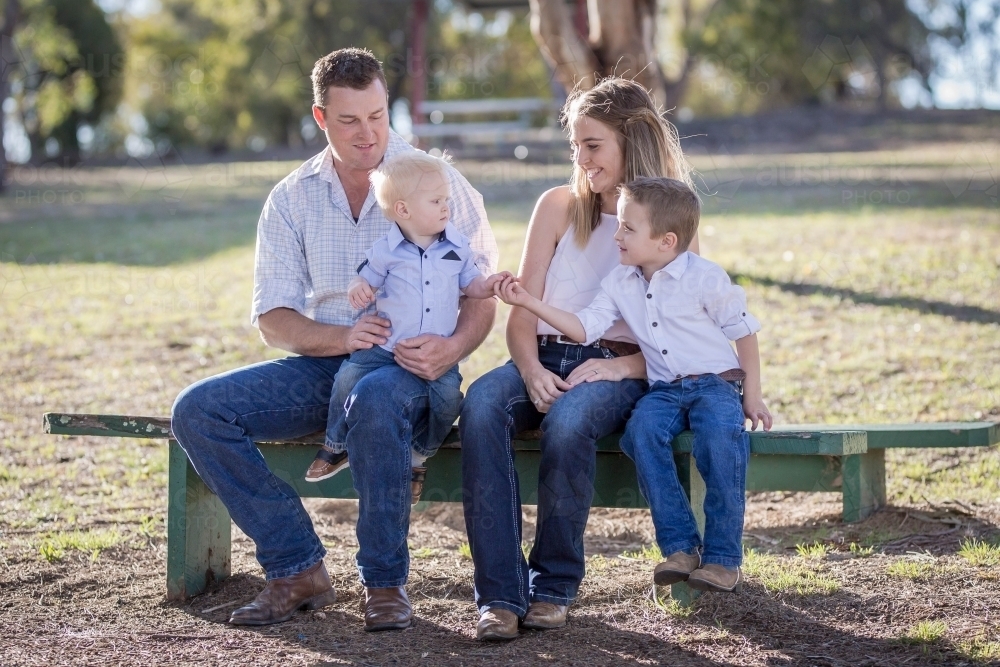 Family sitting together on bench with big brother holding baby brother's hand - Australian Stock Image