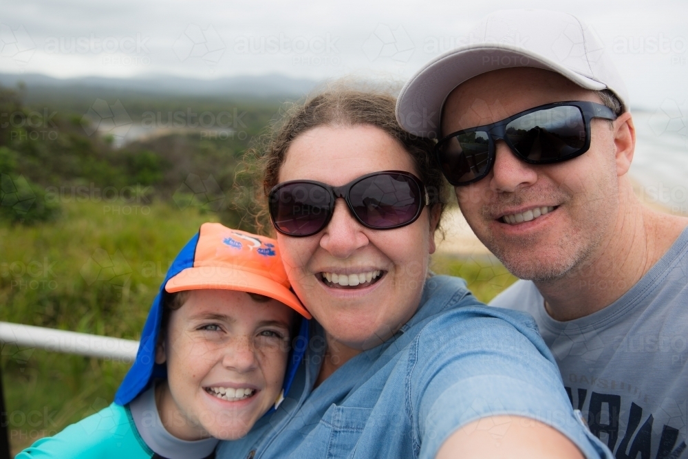 Family standing together happy smiling taking selfie with beach and trees behind - Australian Stock Image