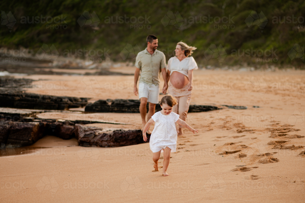 Family strolls along sandy beach at sunset, with a young girl running ahead of her parents - Australian Stock Image