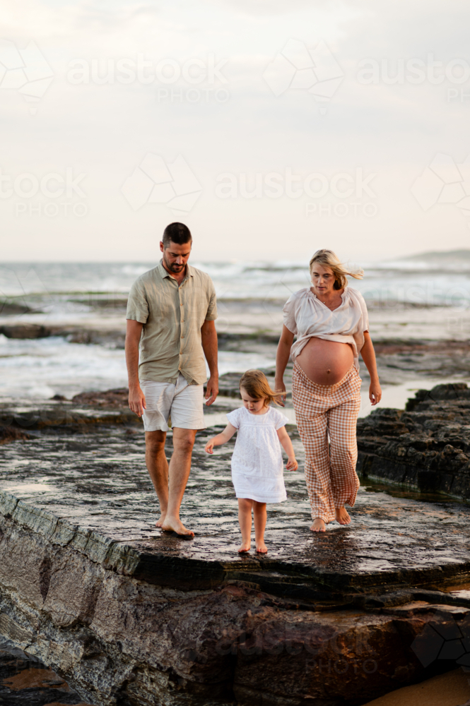 Family walking on rocky beach at sunset with pregnant mother and young daughter - Australian Stock Image