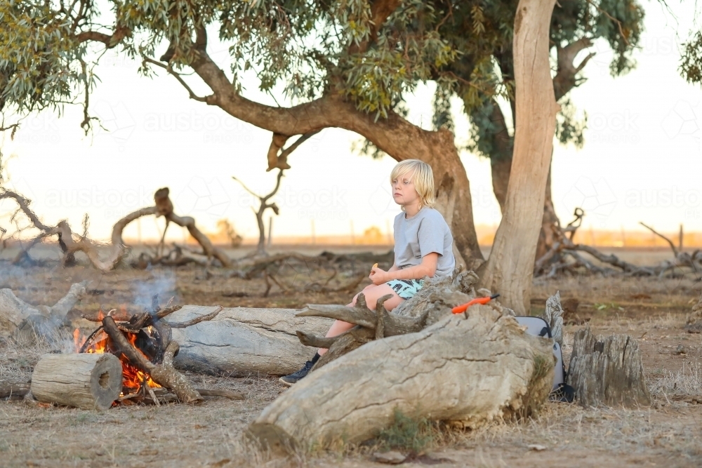 Farm boy sitting on log seat near camp fire in the Australian bush - Australian Stock Image