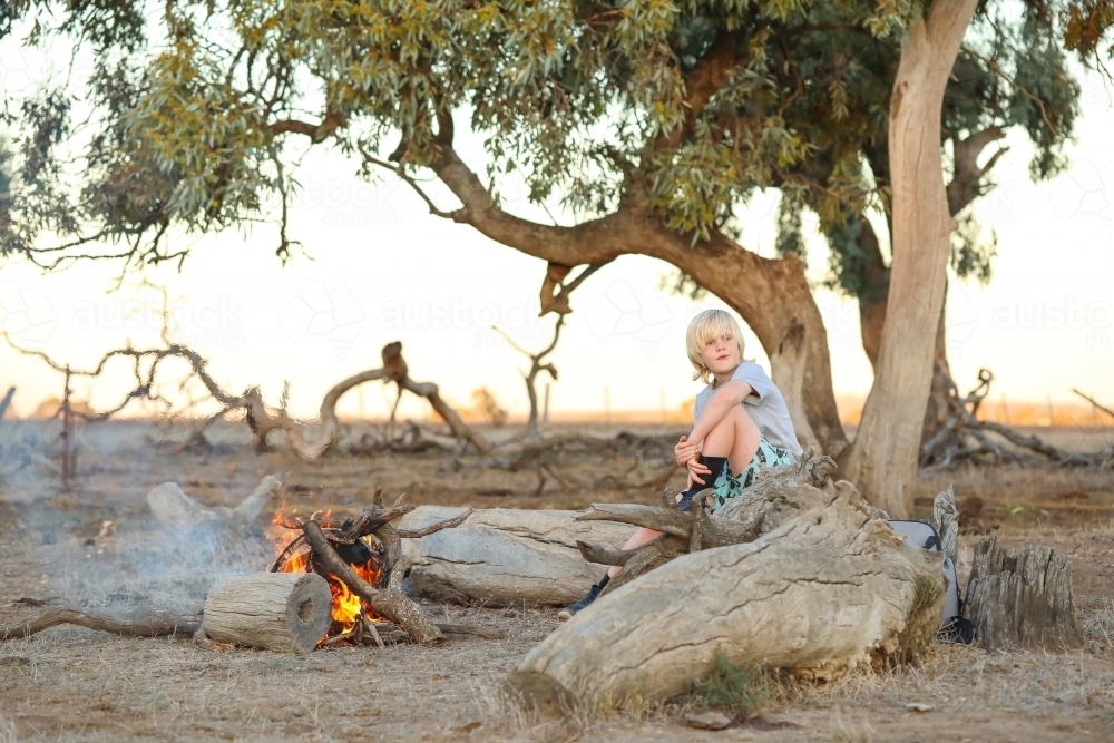 Farm boy sitting on log seat near camp fire in the Australian bush - Australian Stock Image