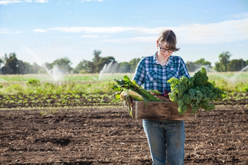Farm girl with glasses in paddock holding box of raw veggies and produce grown on land - Australian Stock Image