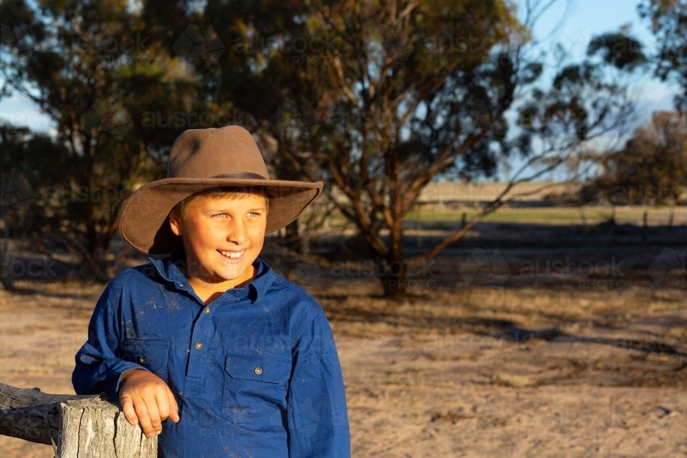 Farm kid wearing work shirt and hat leaning on a fencepost. - Australian Stock Image