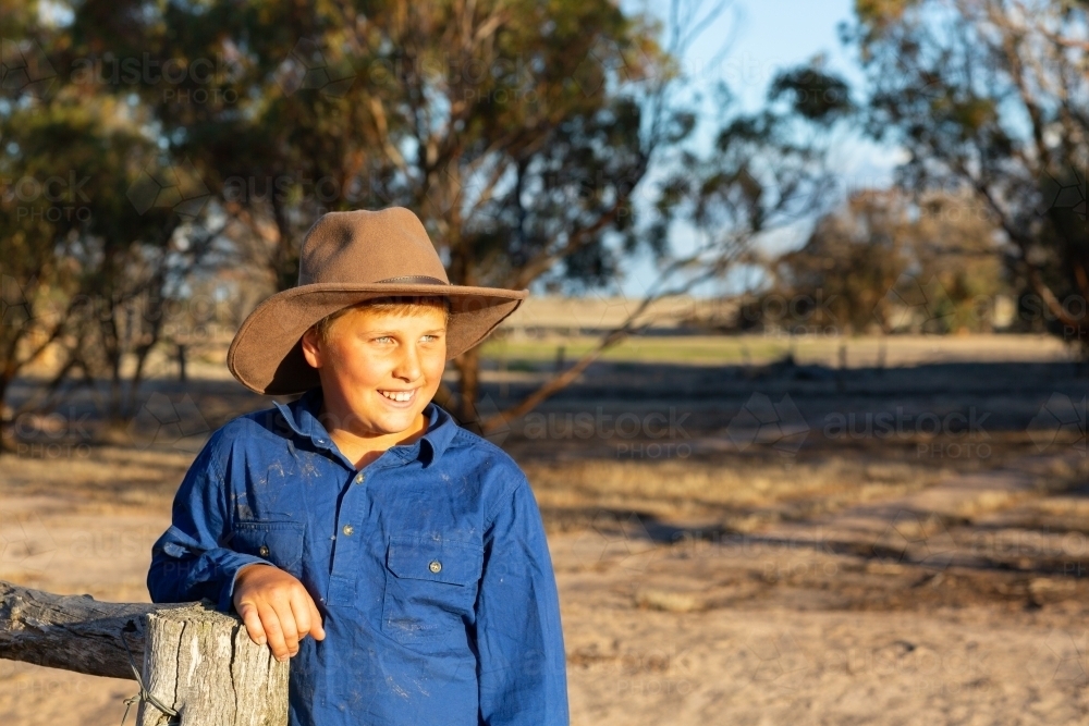Farm kid wearing work shirt and hat leaning on a fencepost. - Australian Stock Image