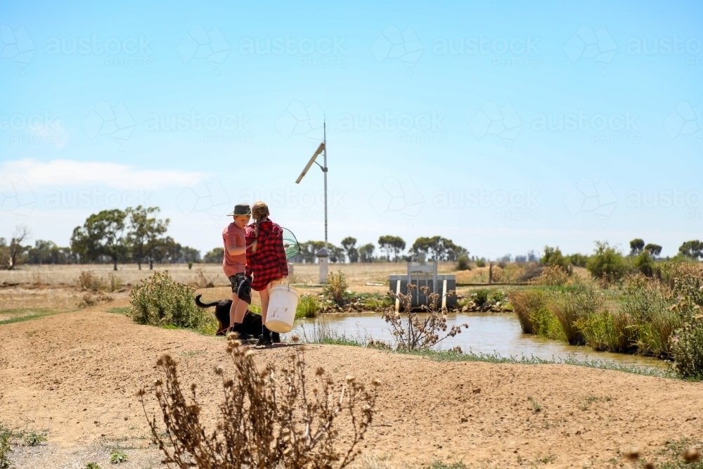 Farm kids standing on channel bank with bucket to catch yabbies - Australian Stock Image