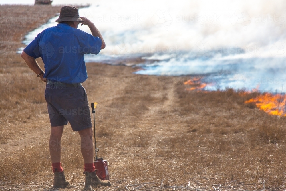 Farmer standing next to a fire lighter watching a fire burning grass - Australian Stock Image