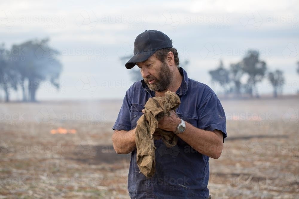 Farmer wiping hands on dirty cloth with burning stubble in background - Australian Stock Image