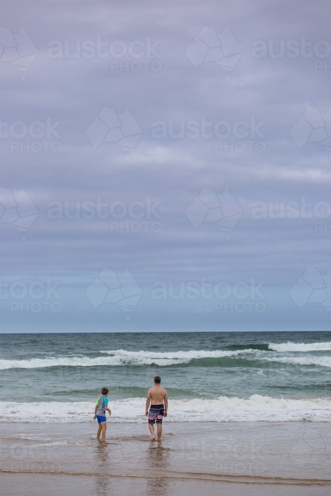 Father and son from behind standing in water on beach looking at waves - Australian Stock Image