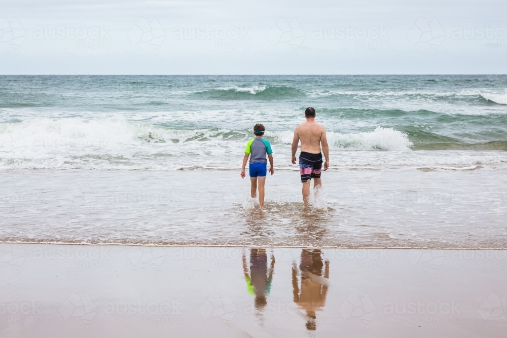 Father and son from behind walking into waves on beach - Australian Stock Image