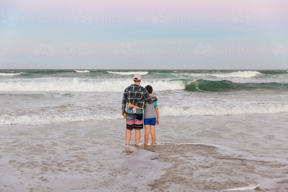 Father and son standing arm in arm on beach looking at waves at sunset - Australian Stock Image