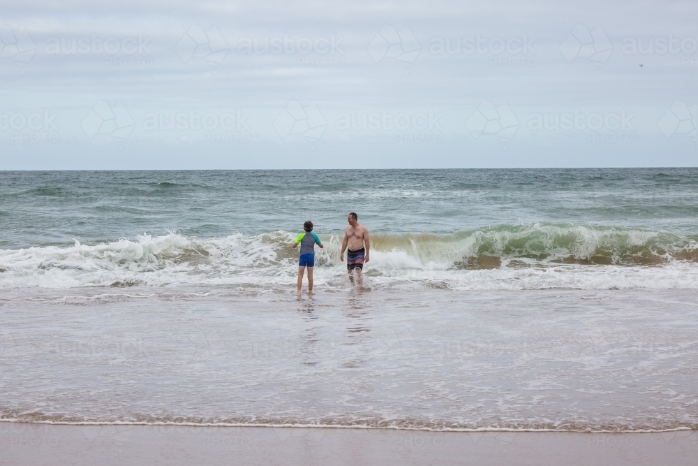 Father and son standing in water on beach looking at waves - Australian Stock Image