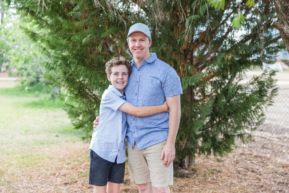 Father and son standing together under tree at home in yard smiling - Australian Stock Image