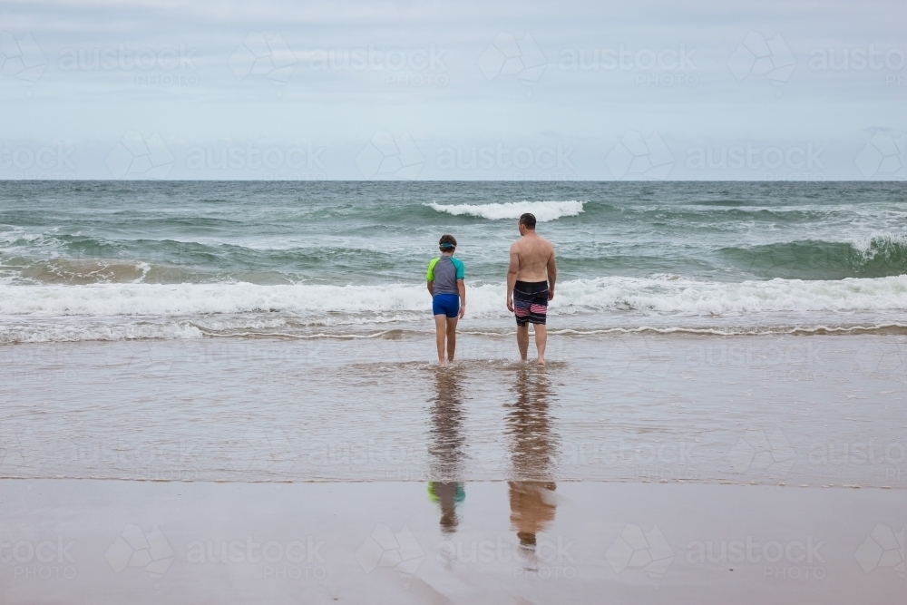 Father and son walking out towards waves at beach from behind - Australian Stock Image