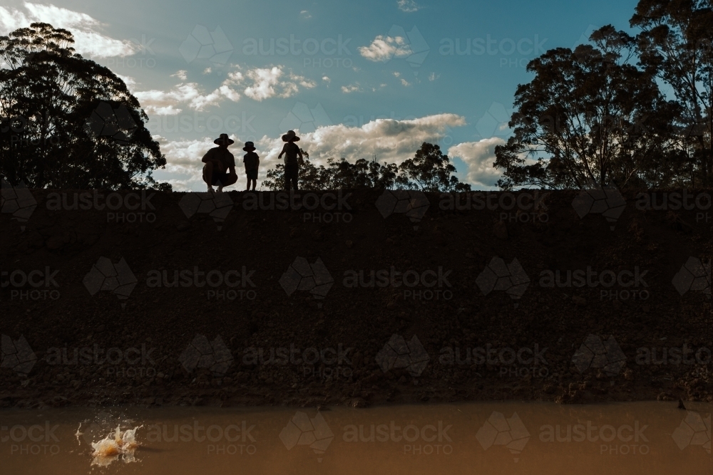 Father and sons throw rock into dam from dam wall - Australian Stock Image