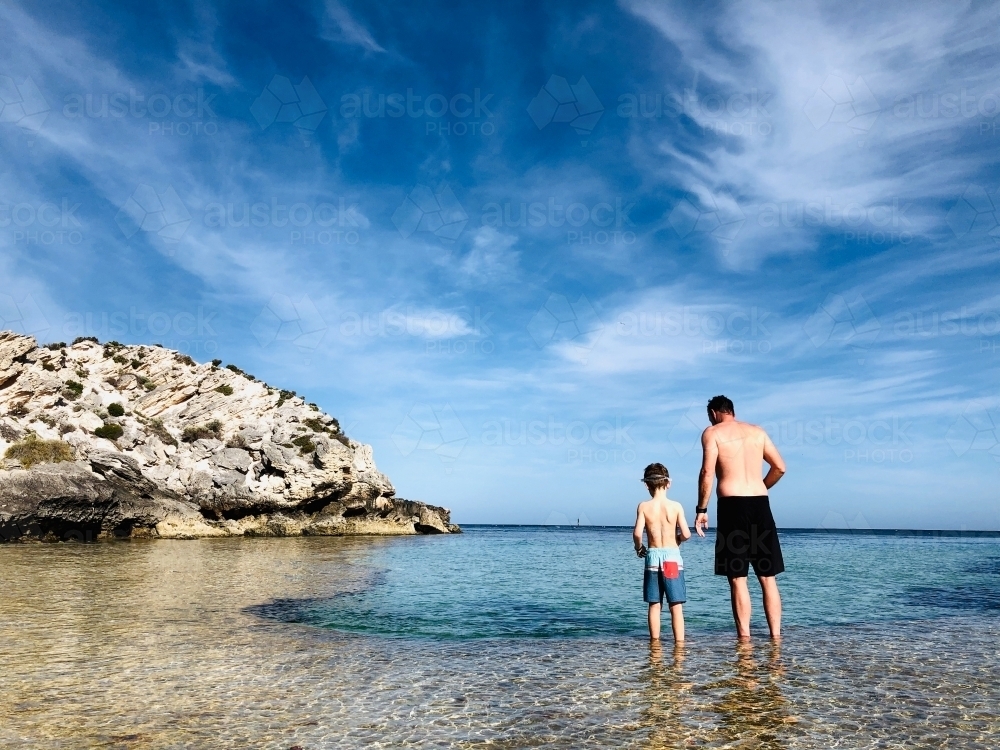 Father and young boy standing on reef overlooking a lagoon wearing board shorts - Australian Stock Image