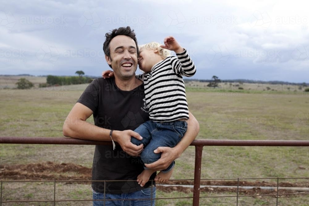 Father and young son laughing together on old gate on rural farm - Australian Stock Image