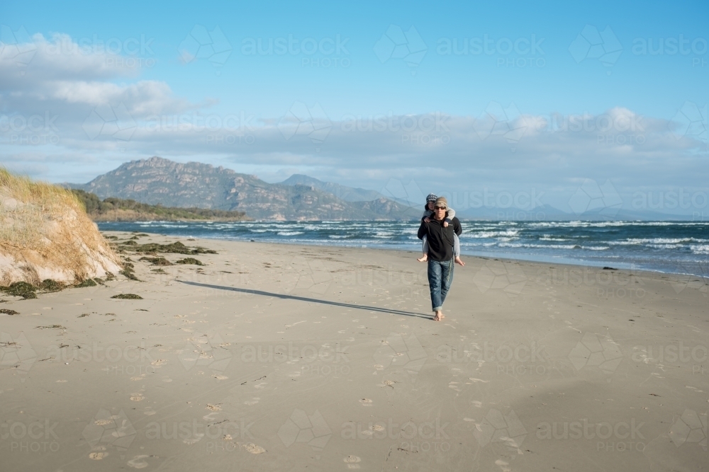 Father carrying son on his back along beach - Australian Stock Image
