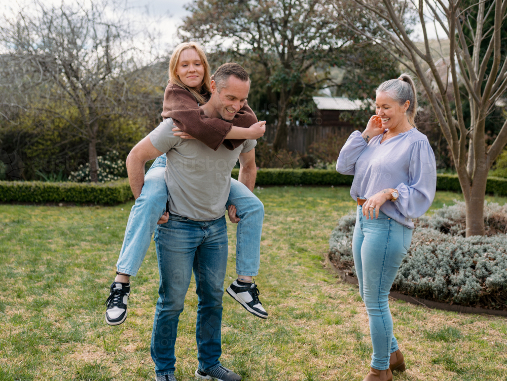Father carrying teenage daughter piggyback while mum watches them in the garden - Australian Stock Image