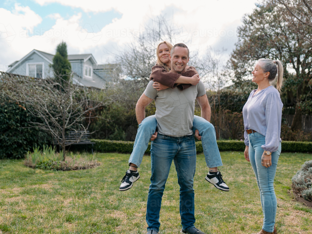 Father carrying teenage daughter piggyback while mum watches them in the garden - Australian Stock Image