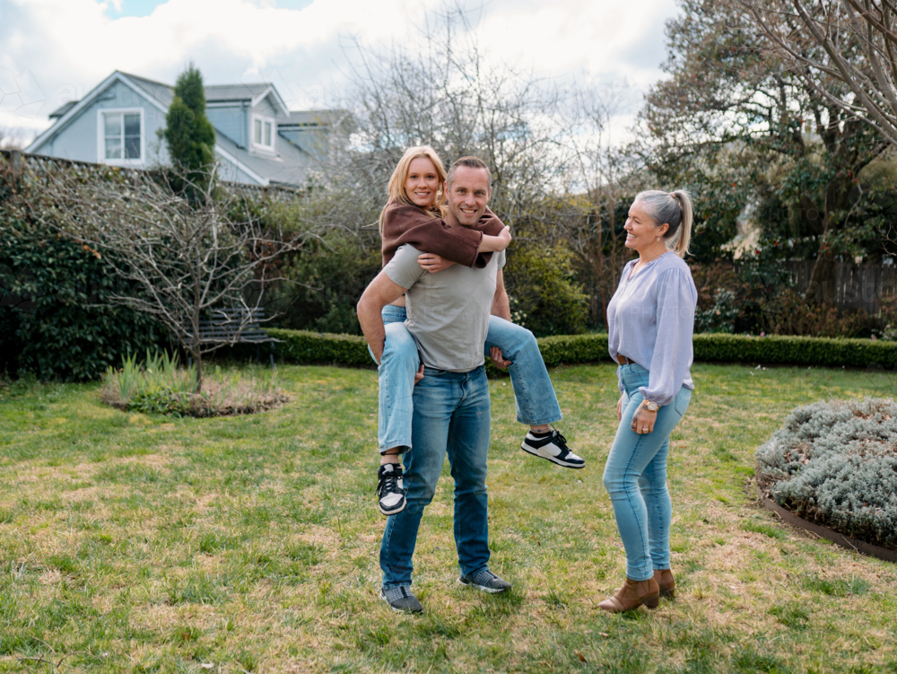 Father carrying teenage daughter piggyback while mum watches them in the garden - Australian Stock Image