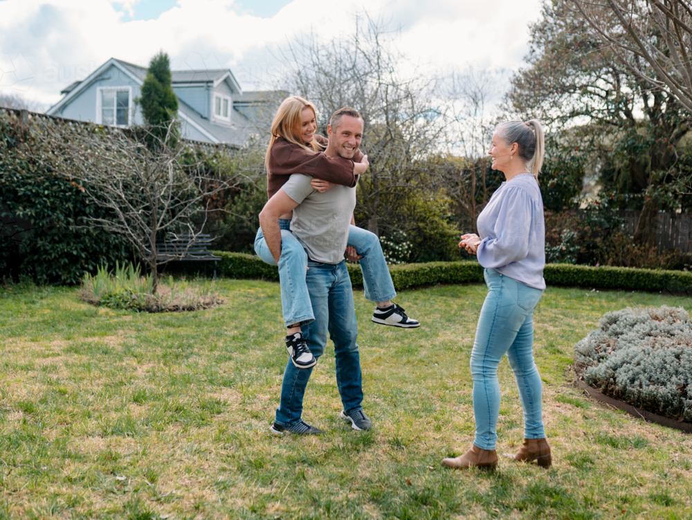 Father carrying teenage daughter piggyback while mum watches them in the garden - Australian Stock Image